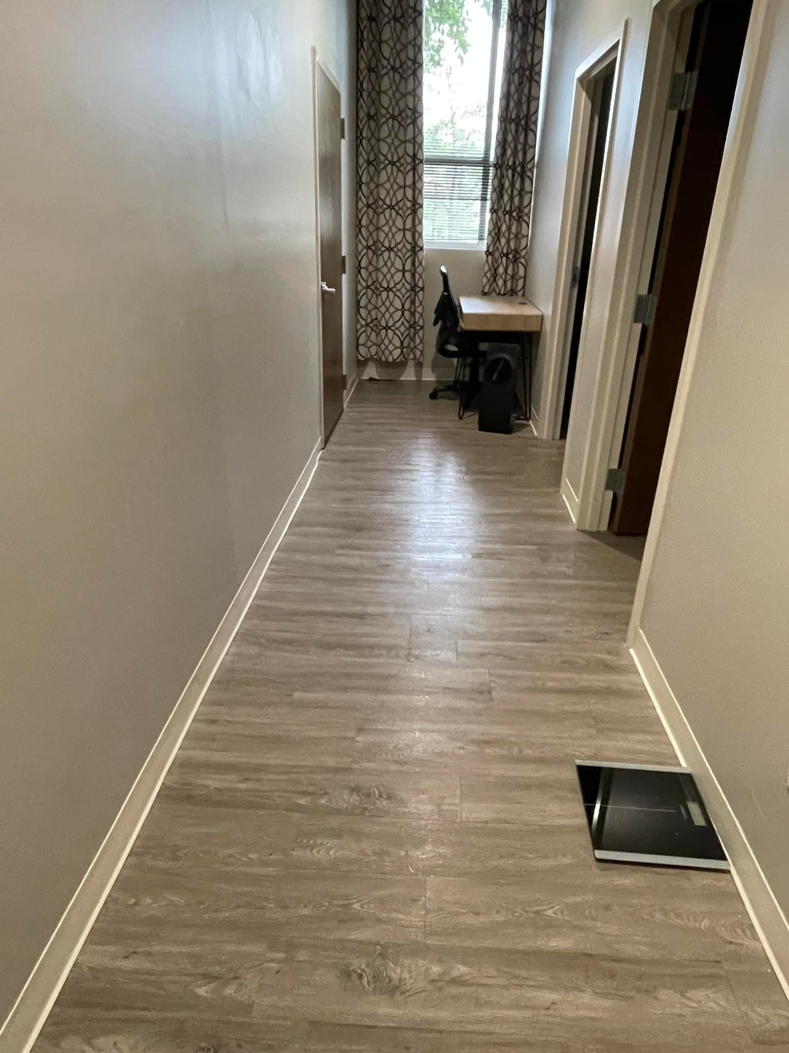 A narrow hallway in a home with light wooden flooring, white walls, and a dark chair visible at the end near a window with curtains. there's an open door on the right and a black object on the floor.
