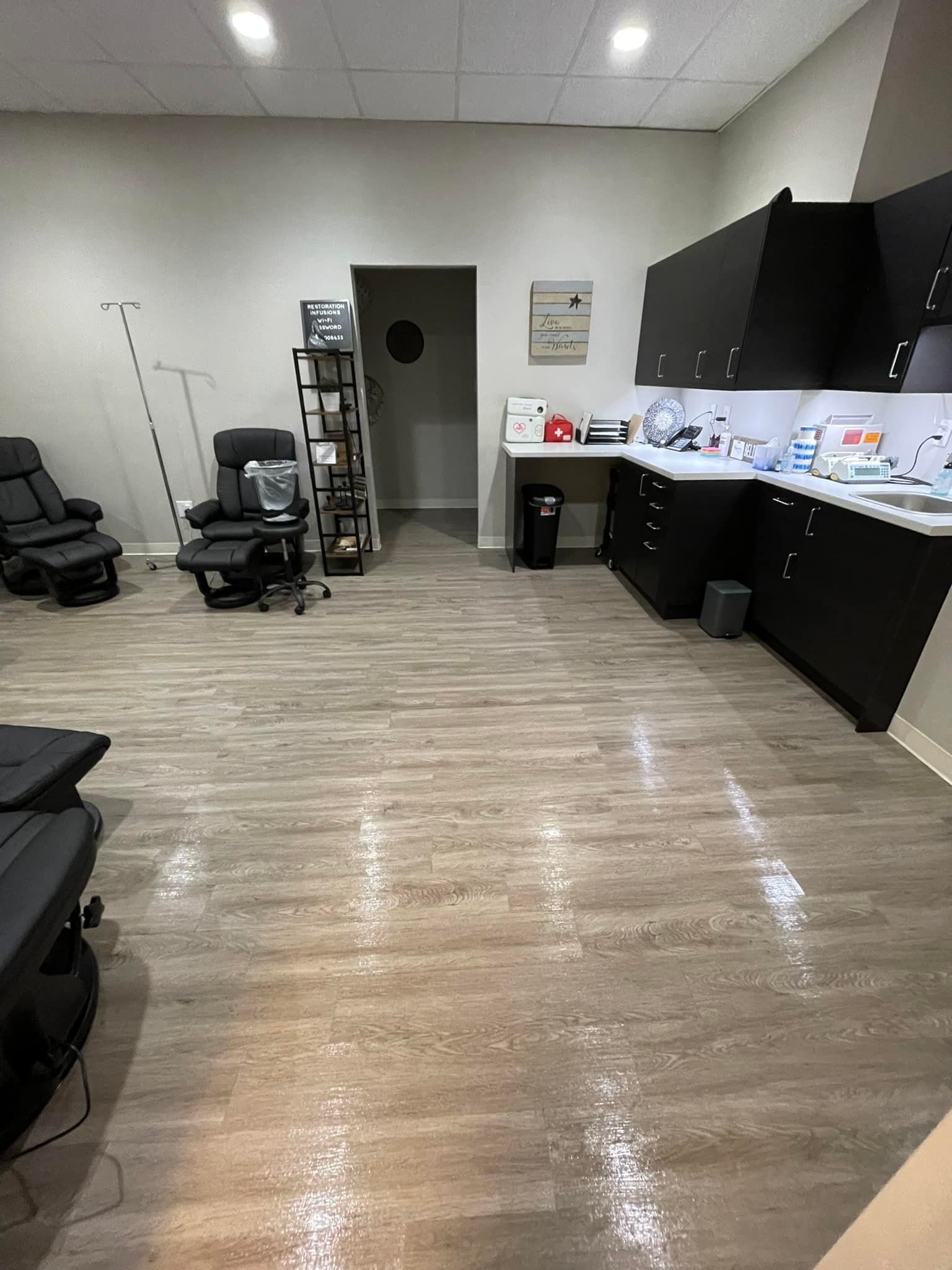 A clean and organized medical office with two reclining chairs, a shelving unit with supplies, a sink, cabinets, and a small trash can on a wooden floor.
